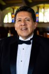 A picture of Ricardo "Ric" Campero wearing a black suit jacket, white dress shirt, and black bow tie smiling with stained glass windows behind him.