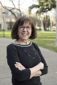 A photo of Joan Calonico wearing glasses and smiling with her arms crossed wearing a black sweater 3/4 sleeve sweater and a necklace.
